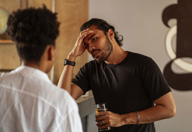 man talking to a woman with his right hand on his forehead with a glass of beer on his left hand
