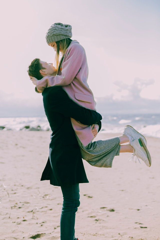 man in gray coat carrying a woman wearing pink coat near the beach 