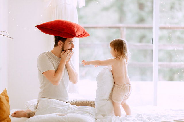 man with red pillow on his head with his kid on bed 