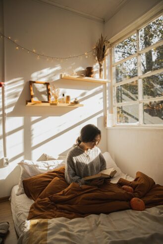 woman in gray long sleeve shirt sitting on her bed reading a book